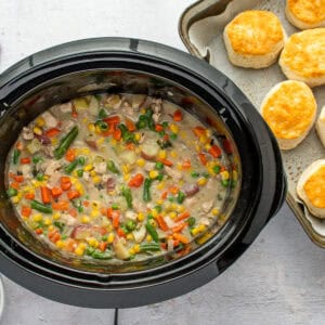 A slow cooker filled with chicken and veggie filling, next to a tray of biscuits.