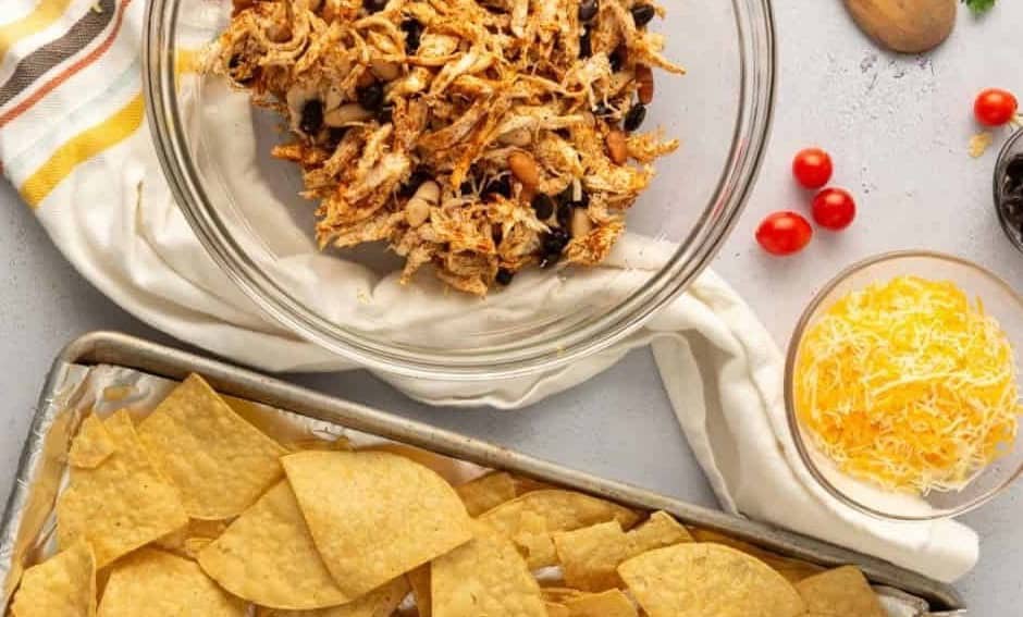 Mexican chicken and bean filling in a bowl, next to a tray of tortilla chips, and a bowl of shredded cheese.