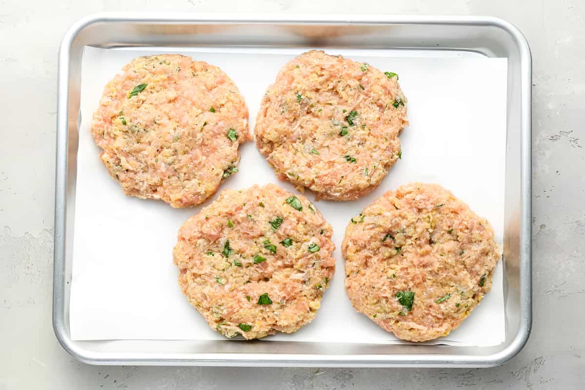 4 raw ground chicken burger patties on a baking sheet.