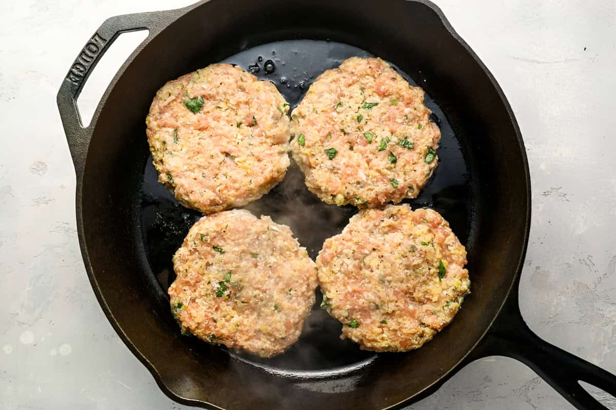 cooking 4 raw ground chicken burger patties in a cast iron pan.