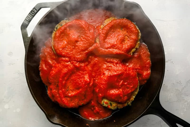 four chicken burger patties topped with marinara sauce in a cast iron pan.