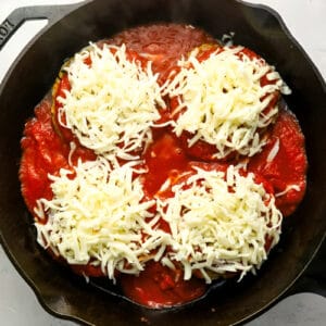 four chicken burger patties topped with marinara and shredded cheese in a cast iron pan.