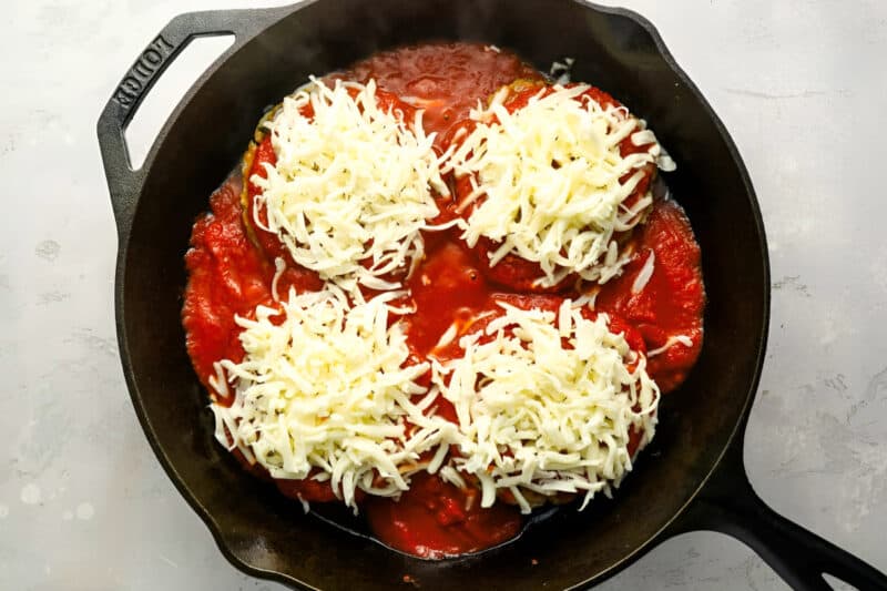 four chicken burger patties topped with marinara and shredded cheese in a cast iron pan.