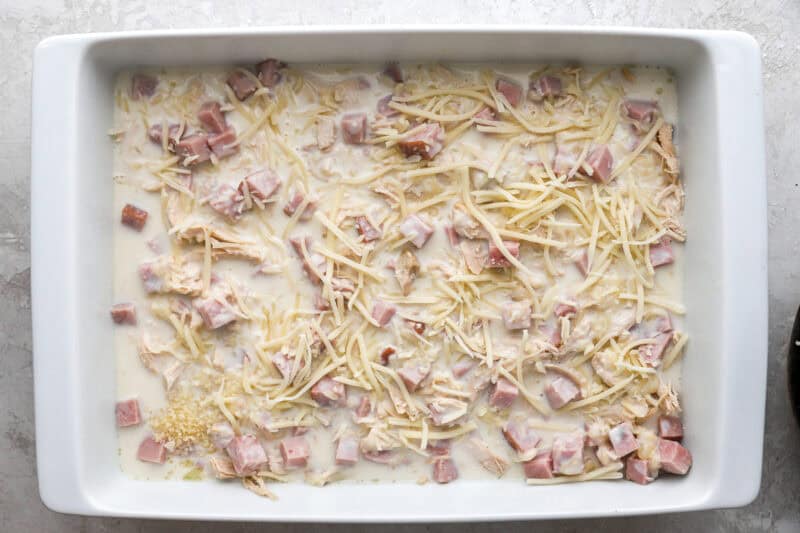 layers of shredded chicken, cubed ham, white sauce, and cheese in a casserole dish.