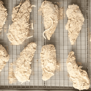 Uncooked chicken strips coated in flour breading mixture, lined up on a baking tray.