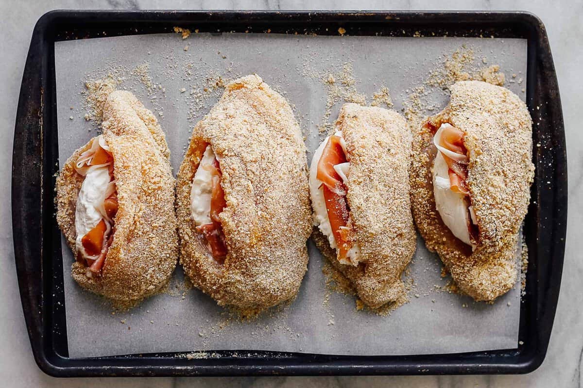 Uncooked chicken breasts stuffed with prosciutto and cheese, on a baking sheet.