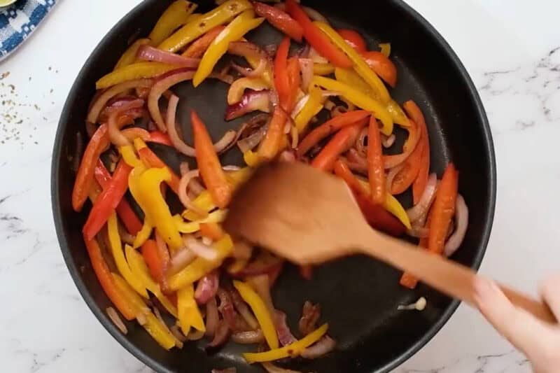 sauteeing sliced peppers and onions in a skillet with a wooden spatula.