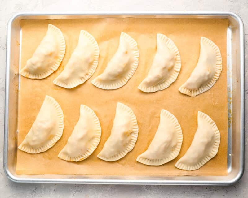 10 uncooked empanadas on a parchment-lined baking sheet.