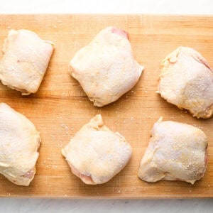 six salted raw chicken thighs on a wooden cutting board.