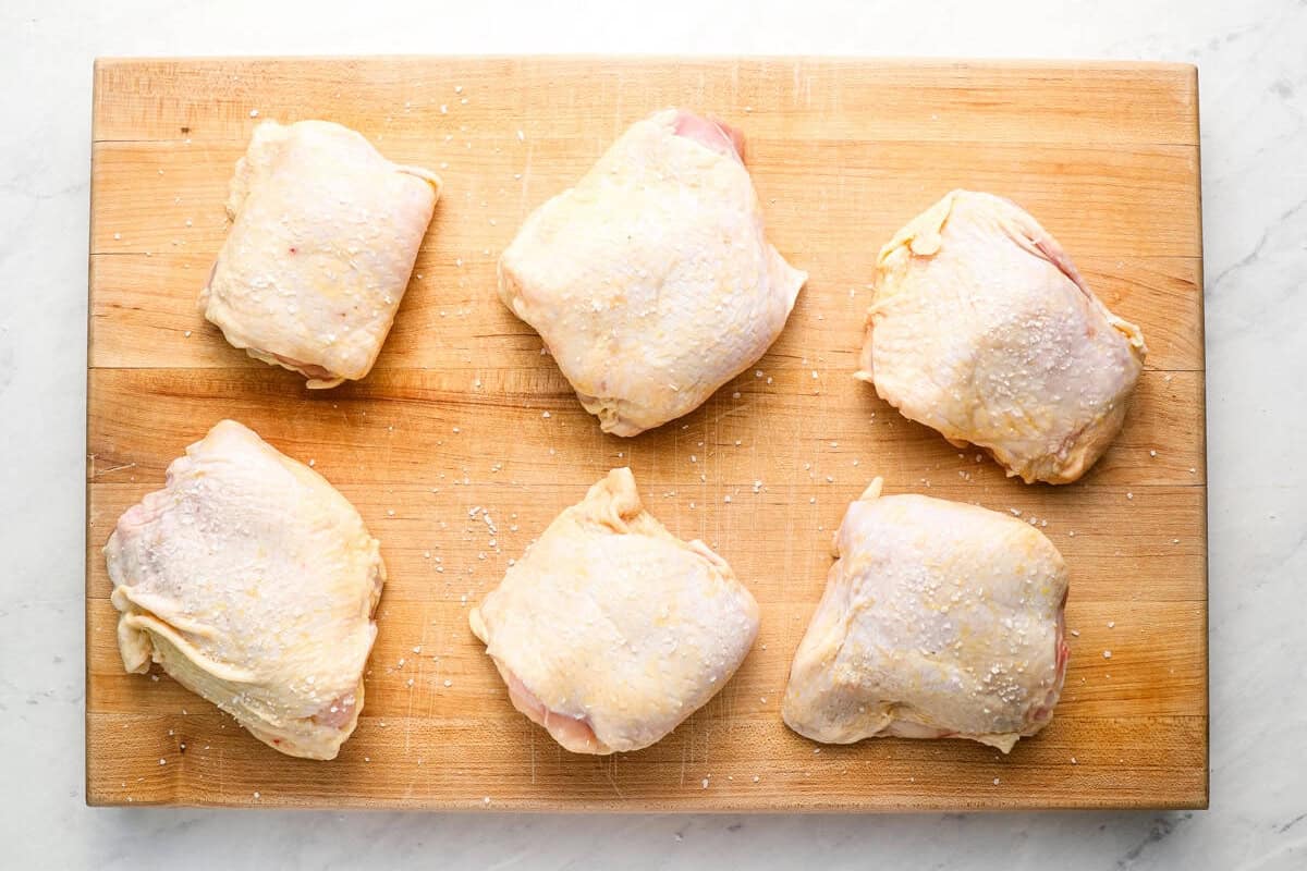 six salted raw chicken thighs on a wooden cutting board.