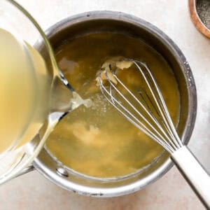 pouring chicken broth over a roux in a saucepan with a whisk.