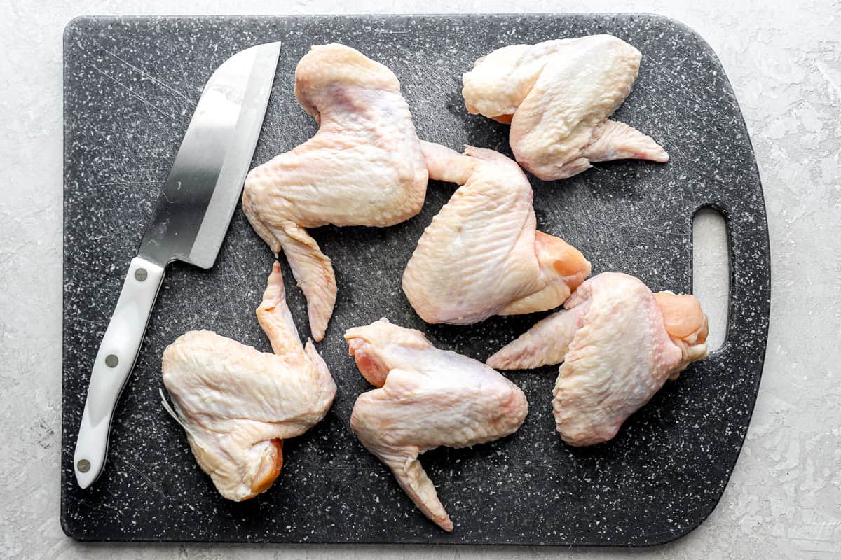 6 whole raw chicken wings on a cutting board with a sharp knife.