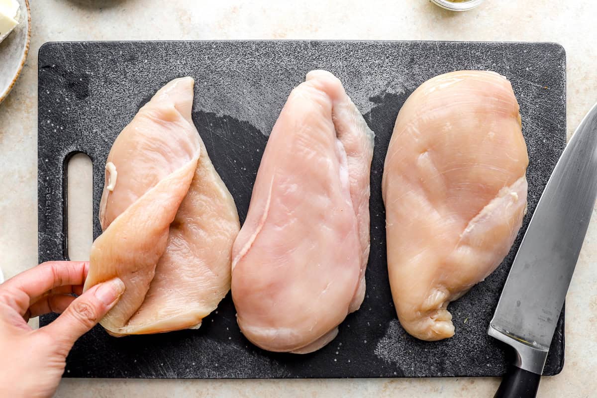 opening a pocket in a raw chicken breast on a cutting board.