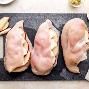 three raw chicken breasts stuffed with apple slices and brie cheese on a cutting board.
