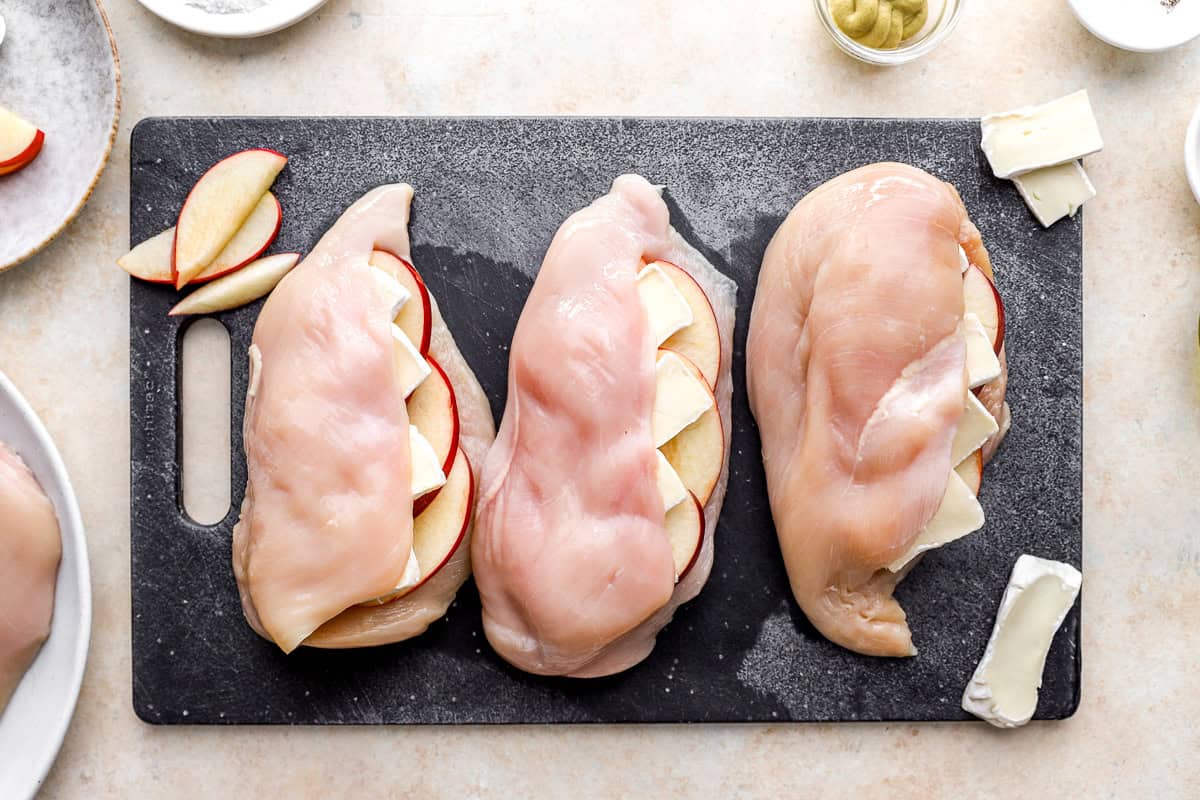 three raw chicken breasts stuffed with apple slices and brie cheese on a cutting board.