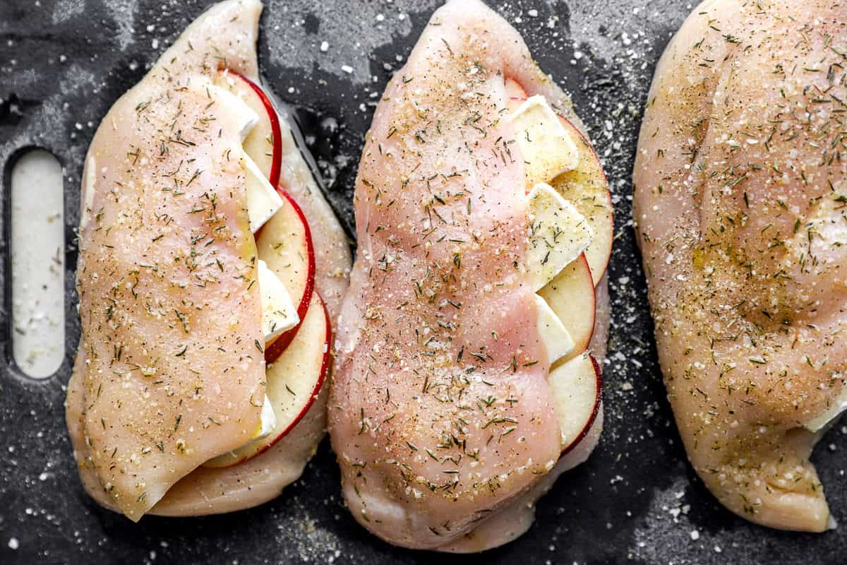 closeup of seasoned apple brie stuffed raw chicken breasts on a cutting board.