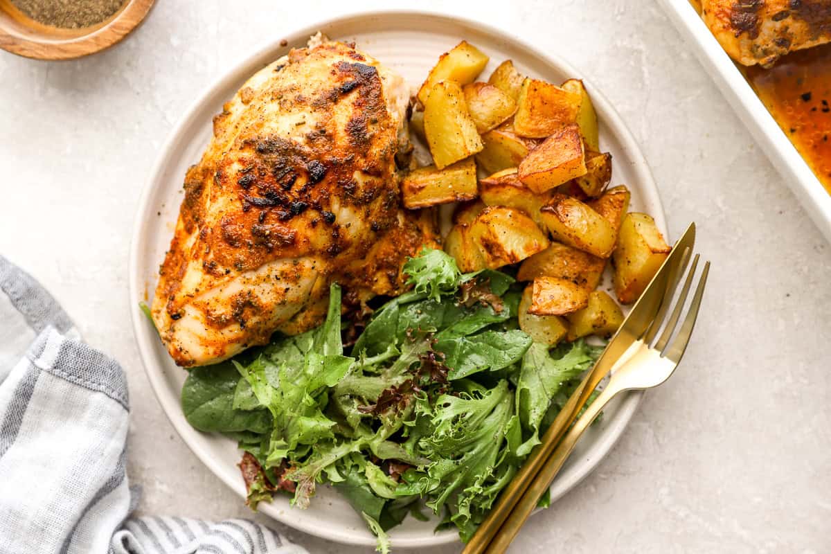 a serving of baked bone in chicken breast on a white plate with salad greens and potatoes.