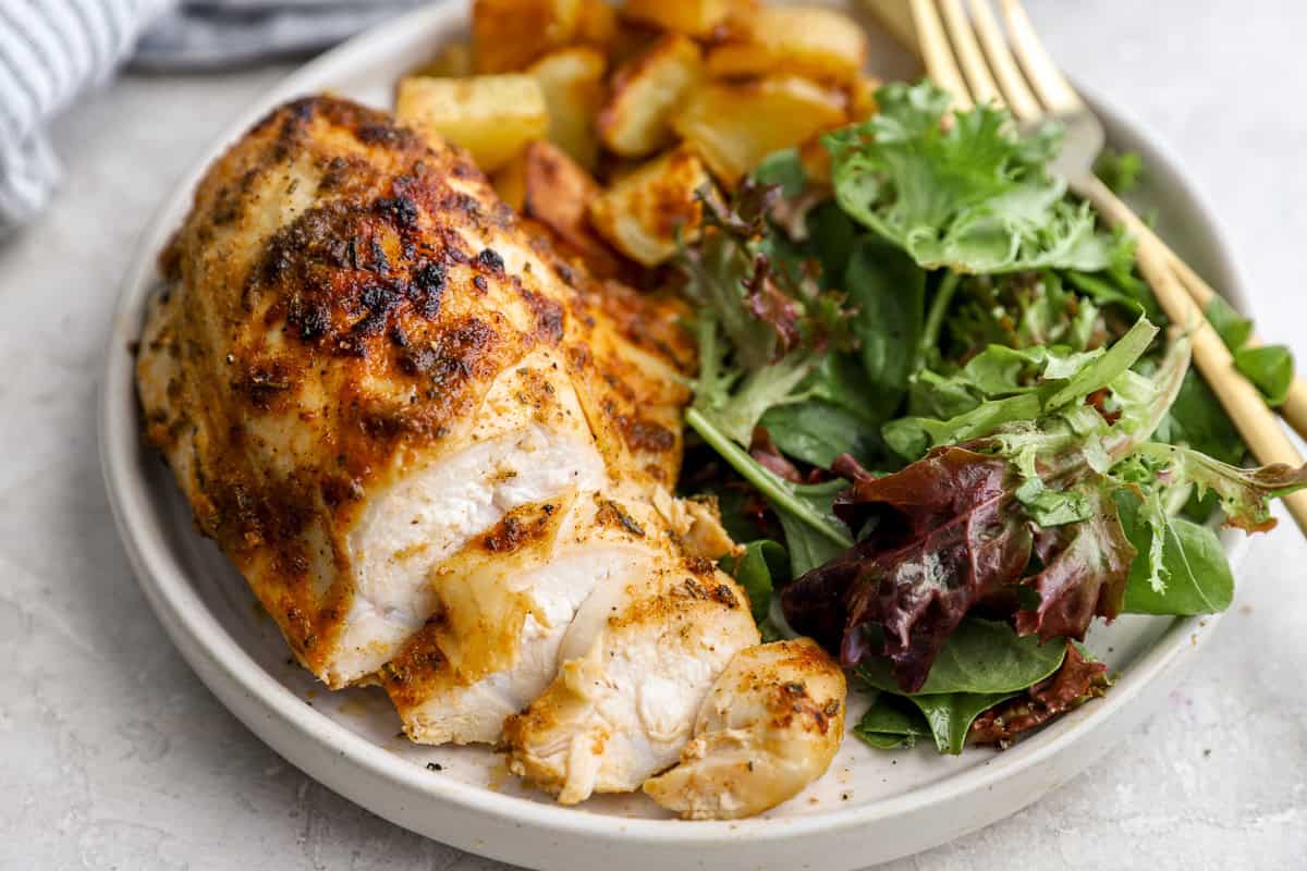 a sliced bone in chicken breast on a white plate with salad greens and potatoes.