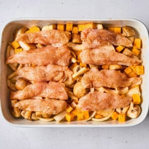 seasoned raw chicken tenders draped over seasoned veggies in a baking dish.
