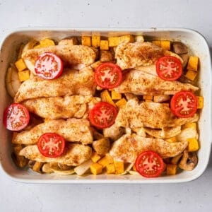 partially cooked chicken tenders and veggies topped with tomato halves in a baking dish.