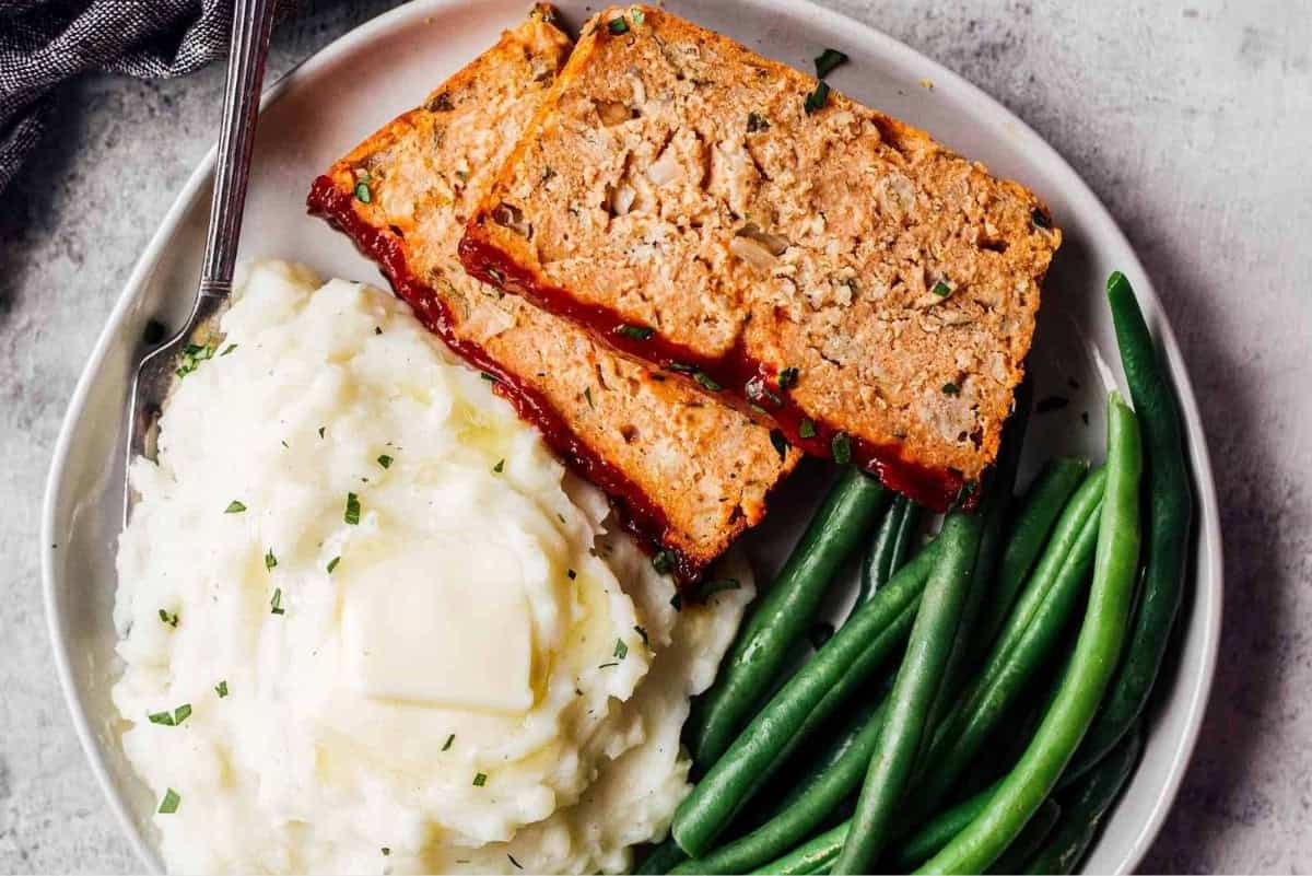 overhead view of 2 slices of chicken meatloaf on a plate with mashed potatoes, green beans, and a fork.