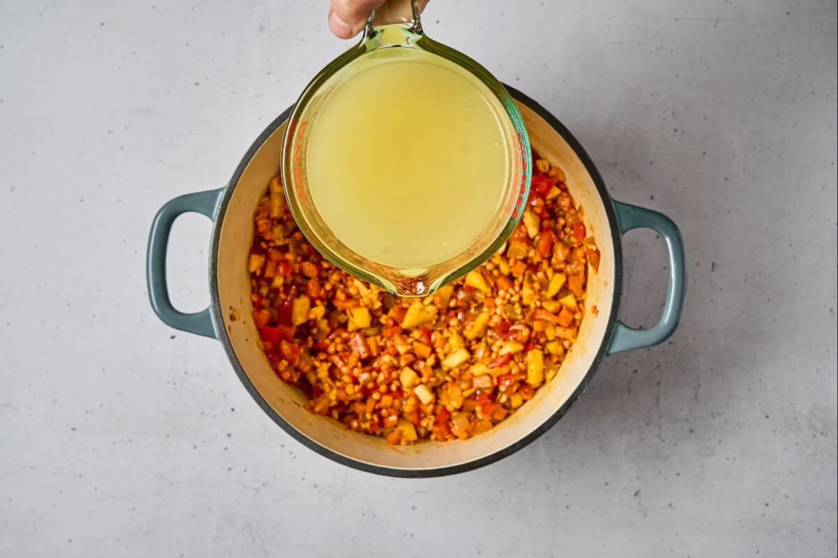 pouring chicken broth over sautéed spiced vegetables and red lentils in a dutch oven.