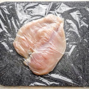 a pounded butterflied chicken breast on a cutting board under plastic wrap.