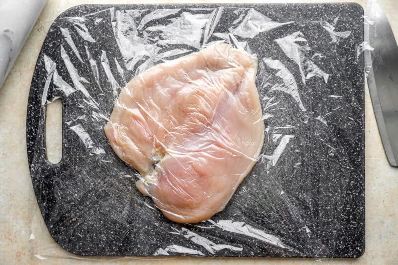 a pounded butterflied chicken breast on a cutting board under plastic wrap.