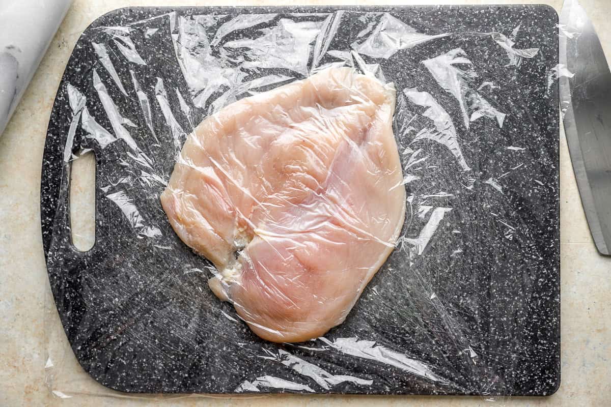 a pounded butterflied chicken breast on a cutting board under plastic wrap.