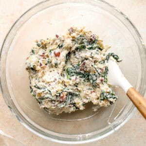 chicken roulade filling with cream cheese, onions, spinach, bacon, and Parmesan in a glass bowl with a rubber spatula.
