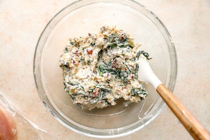 chicken roulade filling with cream cheese, onions, spinach, bacon, and Parmesan in a glass bowl with a rubber spatula.