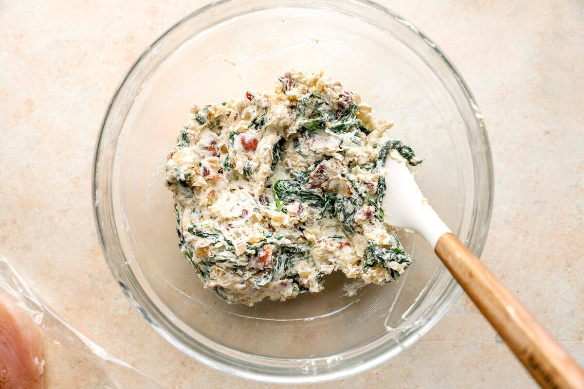 chicken roulade filling with cream cheese, onions, spinach, bacon, and Parmesan in a glass bowl with a rubber spatula.