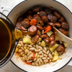 pouring beef broth over chicken sausage rounds, veggies, potatoes, and white beans in a pot.