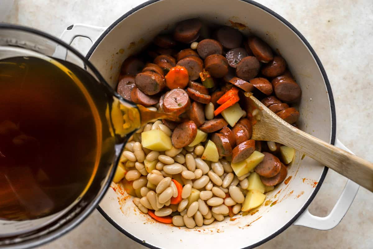 pouring beef broth over chicken sausage rounds, veggies, potatoes, and white beans in a pot.