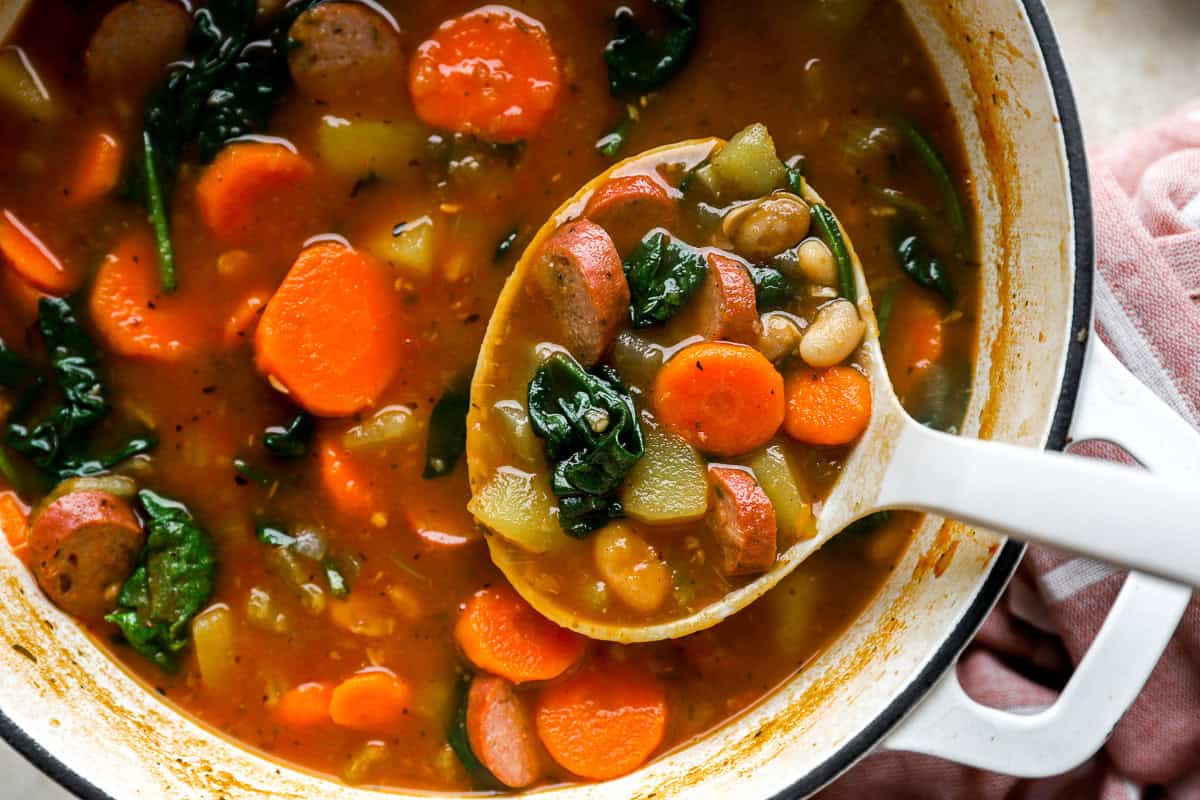 cropped overhead view of scooping a ladleful of chicken sausage white bean soup from a pot.