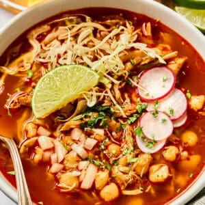 a bowl of chicken pozole rojo topped with sliced radishes, cabbage, cilantro, and a lime wedge.