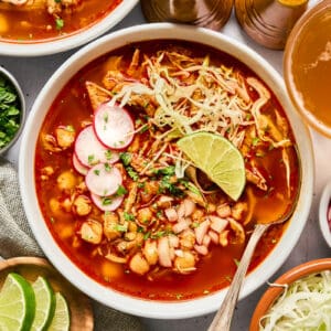 overhead view of a bowl of chicken pozole rojo topped with sliced radishes, cabbage, cilantro, and a lime wedge.
