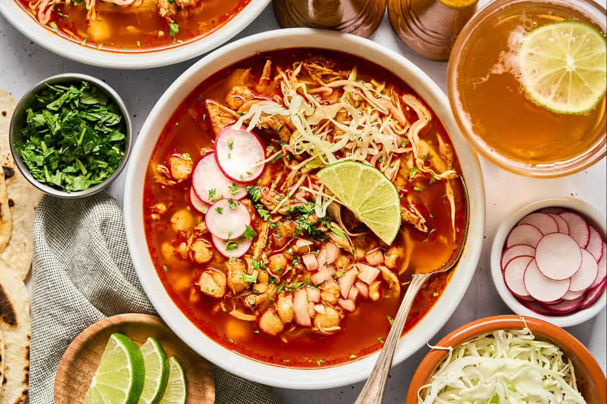 overhead view of a bowl of chicken pozole rojo topped with sliced radishes, cabbage, cilantro, and a lime wedge.