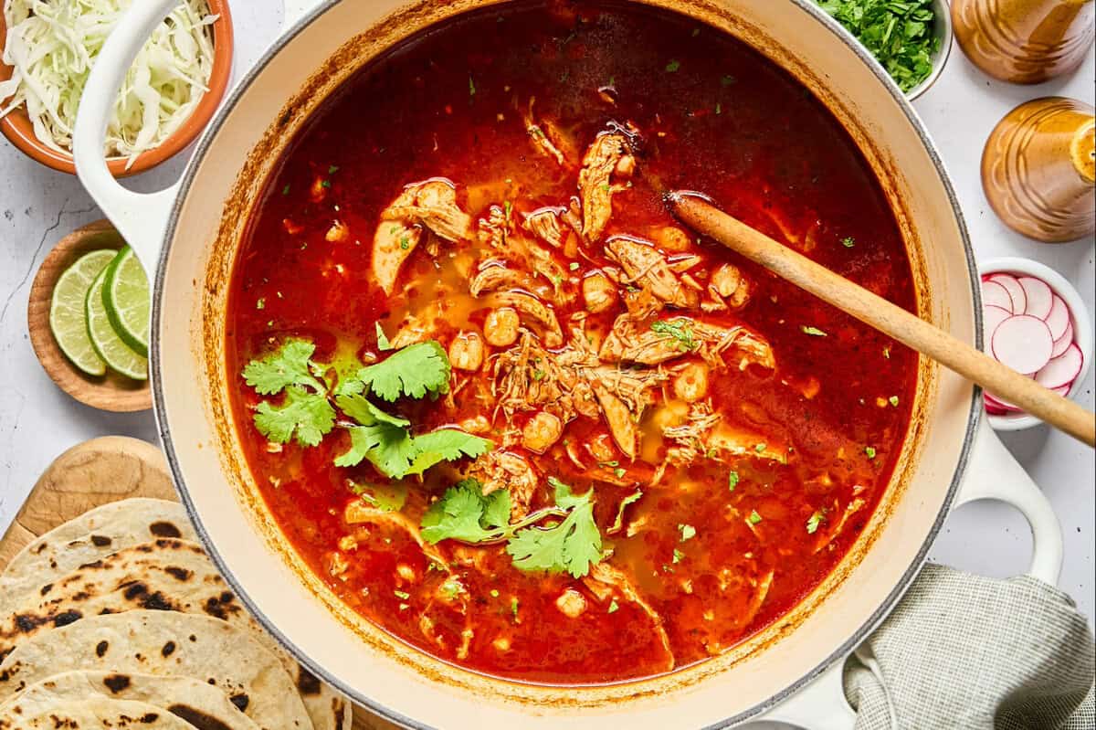 overhead view of chicken pozole rojo in a dutch oven with cilantro and a wooden spoon.