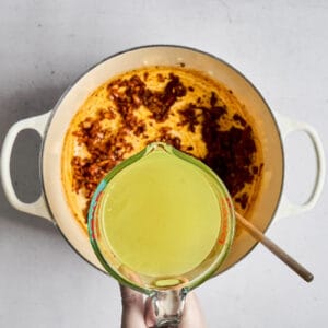pouring chicken broth over sauteed onions, garlic, and spices in a dutch oven.