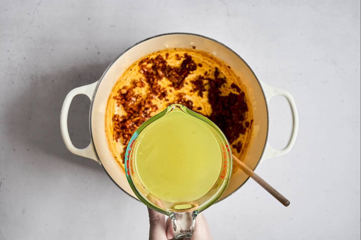 pouring chicken broth over sauteed onions, garlic, and spices in a dutch oven.