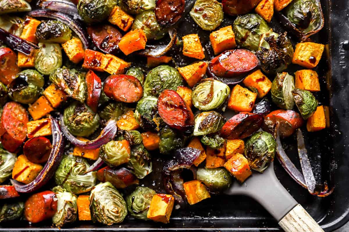 lifting a scoop of sheet pan chicken sausage and veggies from a baking sheet.