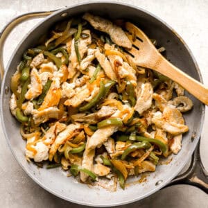 stirring sliced chicken and sautéed peppers and onions together in a pan with a wooden spoon.