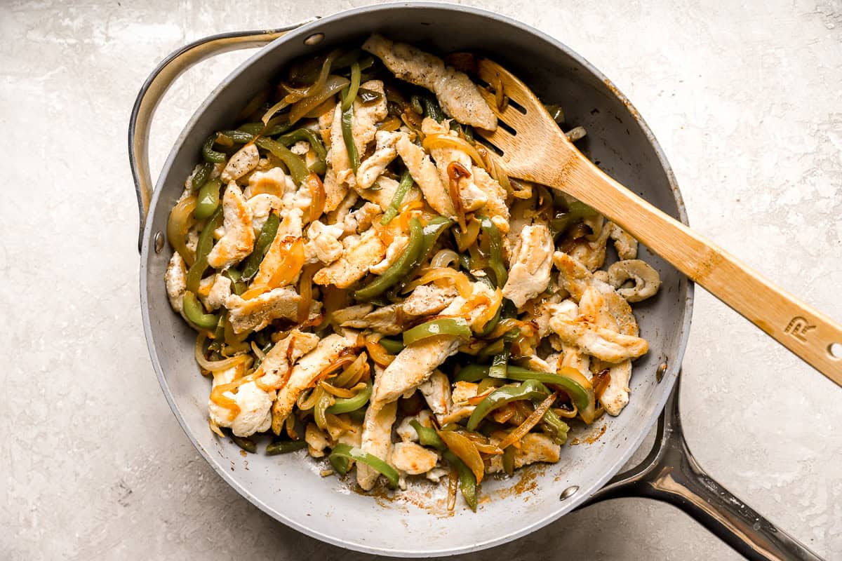 stirring sliced chicken and sautéed peppers and onions together in a pan with a wooden spoon.