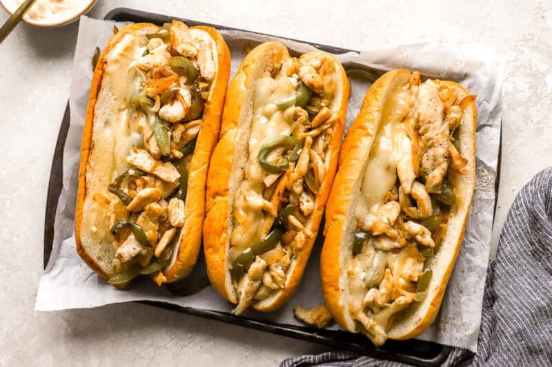 three chicken cheesesteak hoagies on a lined baking sheet.