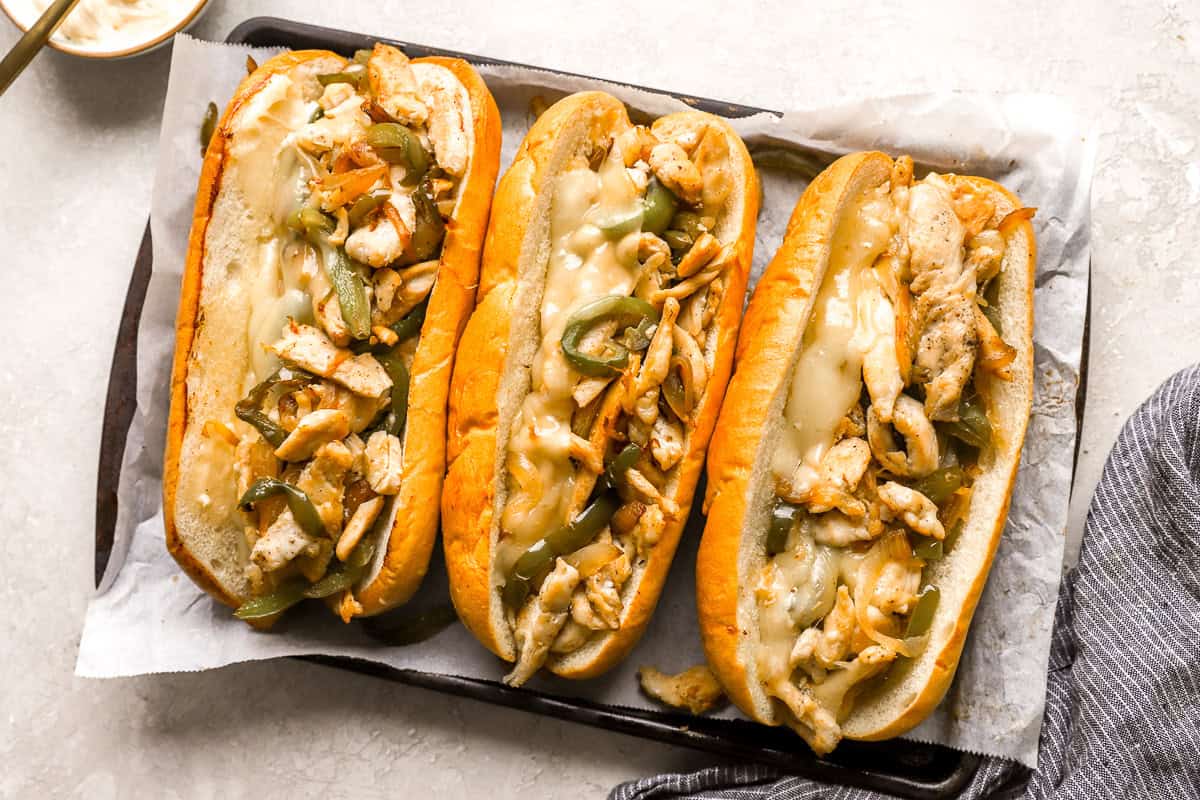 three chicken cheesesteak hoagies on a lined baking sheet.