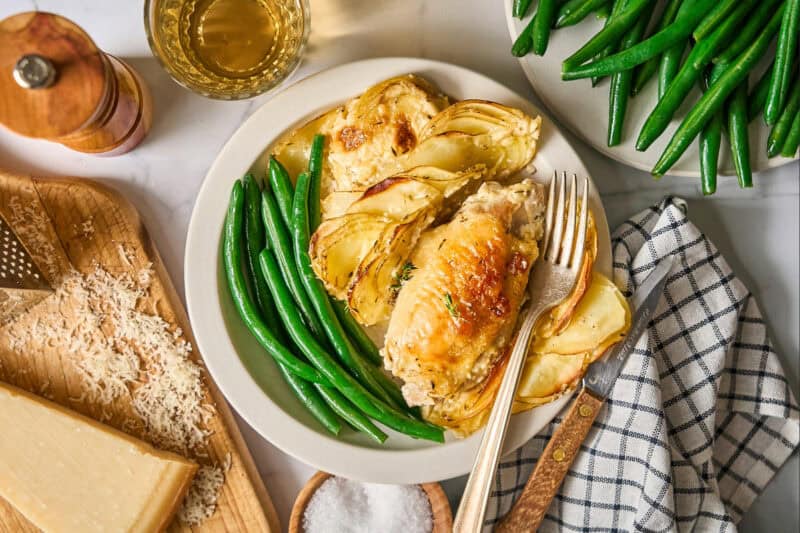 a serving of chicken thighs and potatoes dauphinoise on a white plate with green beans and a fork.