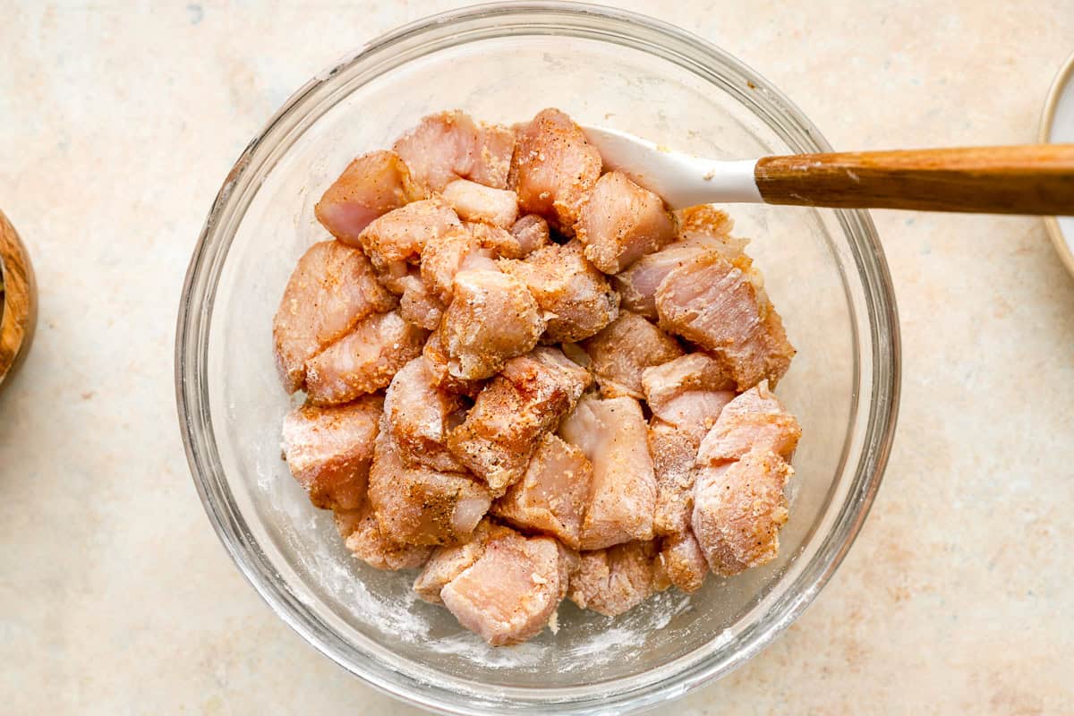 raw chicken bites coated in seasonings in a glass bowl with a rubber spatula.