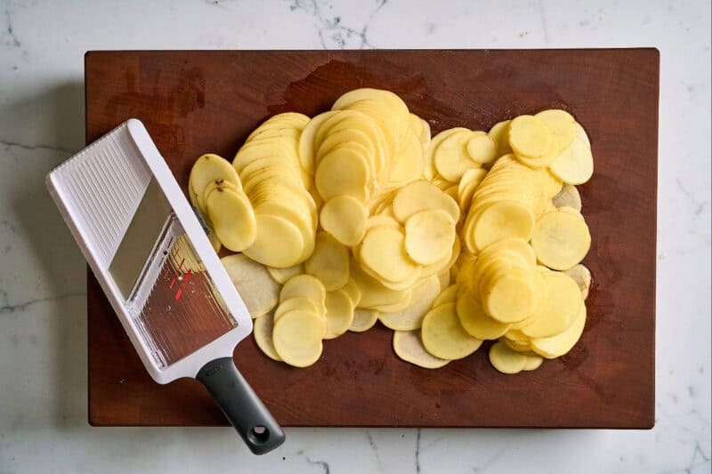 Yukon gold potato slices on a wooden cutting board with a mandoline slicer.