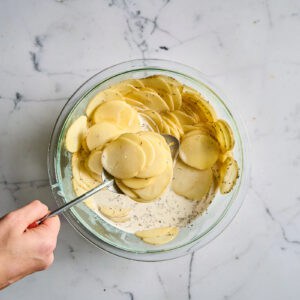 tossing sliced potatoes with seasoned cream and milk in a bowl.